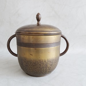 Puede incluir: Un cubo de hielo de latón con tapa y dos asas. El cubo tiene un patrón texturizado en la parte inferior y una banda de líneas elevadas alrededor del centro.