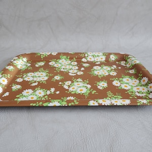Può includere: Un vassoio da portata in metallo marrone con un motivo floreale di margherite bianche. Il vassoio ha un bordo leggermente rialzato.