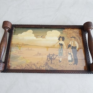 以下が含まれることがあります： 彫刻された取っ手とガラス製のインサートが付いた木製のお盆で、風景の中に家族を描いた絵が描かれています。絵には、フェンスの前に立っている女性、男性、子供と、頭上を飛んでいる鳥が描かれています。背景は薄い茶色で、青い空と白い雲があります。