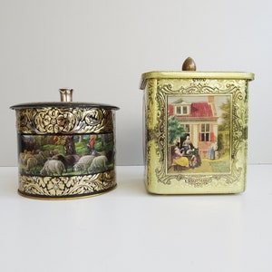 May include: Two vintage metal tins with ornate gold designs. One tin is round and features a black background with a scene of sheep in a field. The other tin is square and has a gold background with a scene of a house and people in a garden.