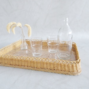 May include: A woven wicker tray with a decorative pattern, holding four clear glass cups and a clear glass bottle. The tray is on a white background.