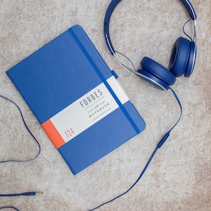May include: A blue notebook with the text "FORBES dotted 240 pages NOTEBOOK" and the number "F24" on the cover. A pair of blue headphones with a cord is lying next to the notebook.