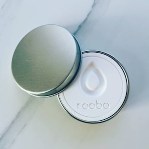 May include: A white round container with a silver lid. The container has a teardrop shape embossed on the top and the word "rooibos" printed on the bottom.