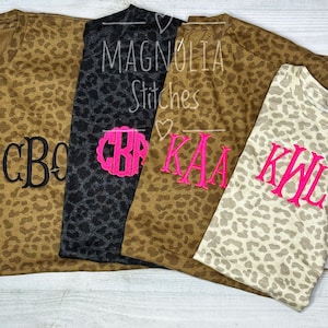 May include: Four leopard print t-shirts with embroidered monograms. The shirts are in shades of brown, gray, and tan. One shirt has a black monogram, one has a pink scalloped monogram, and two have pink monograms. The text "Magnolia Stitches" is visible.