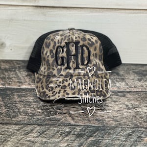 May include: A distressed trucker hat with a leopard print front panel and black mesh back. The front features the embroidered letters "GHD" in black, with the text "Magnolia Stitches" and a heart symbol below.