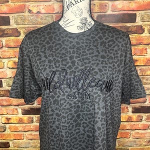 Könnte beinhalten: Ein schwarzes und graues T-Shirt mit Leopardenmuster und dem in Schwarz gestickten Text "Wild Willow Salon".