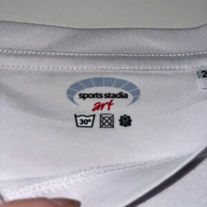 K&ouml;nnte beinhalten: Wei&szlig;es T-Shirt mit dem Logo "sports stadia art" in einem blau-wei&szlig;en Oval. Waschhinweise, einschlie&szlig;lich eines 30&deg;C Waschsymbols, sind sichtbar. Der Stoff scheint weich und atmungsaktiv zu sein.