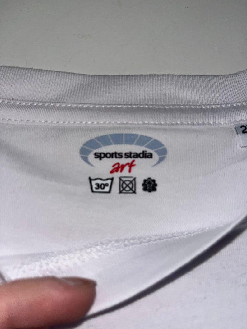 K&ouml;nnte beinhalten: Wei&szlig;es T-Shirt mit dem Logo "sports stadia art" in einem blau-wei&szlig;en Oval. Waschhinweise, einschlie&szlig;lich eines 30&deg;C Waschsymbols, sind sichtbar. Der Stoff scheint weich und atmungsaktiv zu sein.
