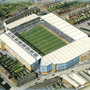 May include: An aerial view of a large, white-roofed stadium with a green soccer field. The stadium is surrounded by a residential neighborhood with houses and a parking lot.