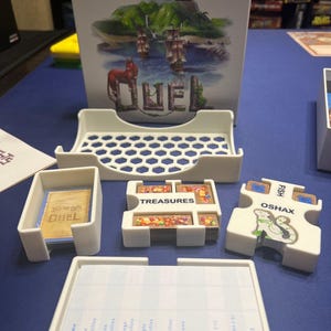 May include: White plastic organiser for the board game "The Isle of Cats: Duel". The organiser has compartments for cards, tokens, and other game components. The game box is in the background.