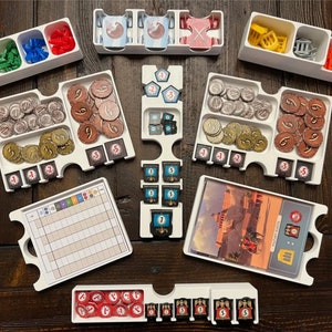 May include: White plastic organizer trays for board game components. The trays contain various colored plastic tokens, dice, and cards. The trays are labeled with numbers and symbols.