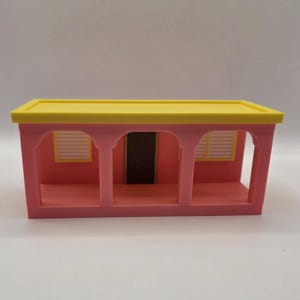 Puede incluir: Una fachada de casa en miniatura con un techo plano de color amarillo brillante y una pared exterior rosa. La estructura incluye tres aberturas arqueadas, dos ventanas blancas con persianas y una puerta rectangular oscura central.