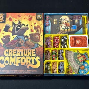 May include: A board game called "Creature Comforts" with a colourful box featuring woodland animals and the text "A game by Roberta Taylor with art by  Graffiti de Tenny". The box is open to reveal a tray with various game pieces, including tokens, tiles, and a fireplace.