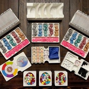 May include: White plastic organizers with compartments for storing game pieces. The organizers contain various colored tokens, dice, and cards. The organizers are arranged on a wooden table.