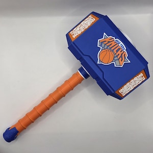 May include: A blue and orange toy hammer featuring the New York Knicks basketball team logo on the side. The hammer head has white decorative patterns on the top and bottom edges, and the handle has a textured grip with a blue end cap.