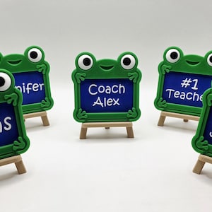 May include: Five green frog-shaped frames with white eyes, each holding a blue rectangular sign with white text. The signs display the names Lucas, Jennifer, Coach Alex, #1 Teacher, and Mr. Johnson. Each frame rests on a small wooden easel stand.