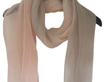 May include: A delicate, lightweight scarf in shades of pale pink and cream, draped over a black hanger. The scarf has a subtle fringe at the ends and appears to be made of a soft, flowing fabric. The color gradient adds visual interest.