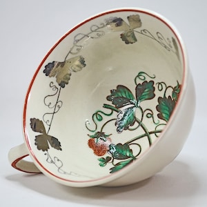 Op de afbeelding: Een witte theekop van keramiek met een rode rand en een bloemenmotief met groene bladeren en rode aardbeien. Het ontwerp bevat gouden accenten.