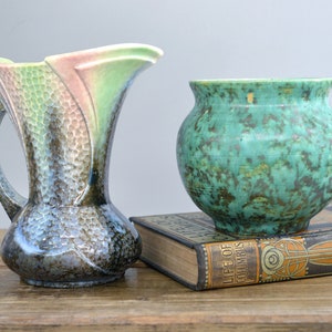 May include: Two vintage ceramic vases, one with a green and brown mottled glaze and the other with a pink, green, and blue iridescent glaze. The vases are sitting on top of a book titled "Life of Columbus".