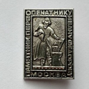 May include: A rectangular silver-colored pin with embossed text and a central figure. The text includes Cyrillic characters and the word "Moscow." The central image depicts a man in historical attire, possibly a printer.