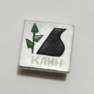 May include: A silver pin with a black and green design. The design features a stylized plant and a black shape. The text "КЛИН" is printed on the pin.