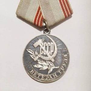 Puede incluir: Una medalla de plata con una estrella roja y una hoz y un martillo, con el texto "CCCP ВЕТЕРАН ТРУДА" alrededor del borde. La medalla está unida a una cinta blanca con dos rayas rojas.