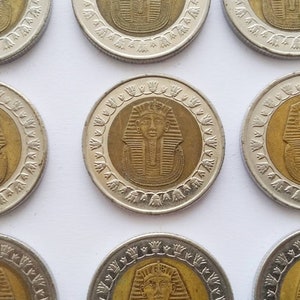 Puede incluir: Nueve monedas egipcias con un centro dorado que representa la cara de un faraón, rodeado por un borde plateado. Las monedas están dispuestas en una cuadrícula de 3x3 sobre una superficie blanca.