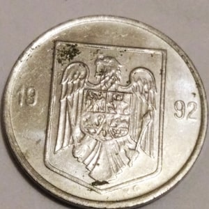 May include: Silver coin with the coat of arms of Romania featuring an eagle with a shield. The coin has the year 1992 inscribed on it.