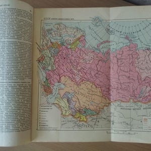 May include: An open book displaying a detailed vintage map of the USSR, with regions colored in pink, yellow, and green. The map is accompanied by text in Russian. The book is open on a wooden surface.