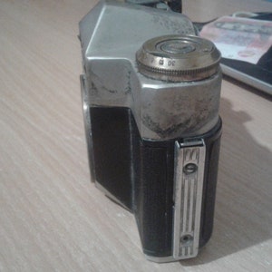 Puede incluir: Una c&aacute;mara de pel&iacute;cula vintage plateada y negra con un acabado desgastado. La c&aacute;mara tiene una cubierta de cuero negro y un cuerpo de metal plateado. La c&aacute;mara tiene un dial en la parte superior con los n&uacute;meros del 1 al 30.