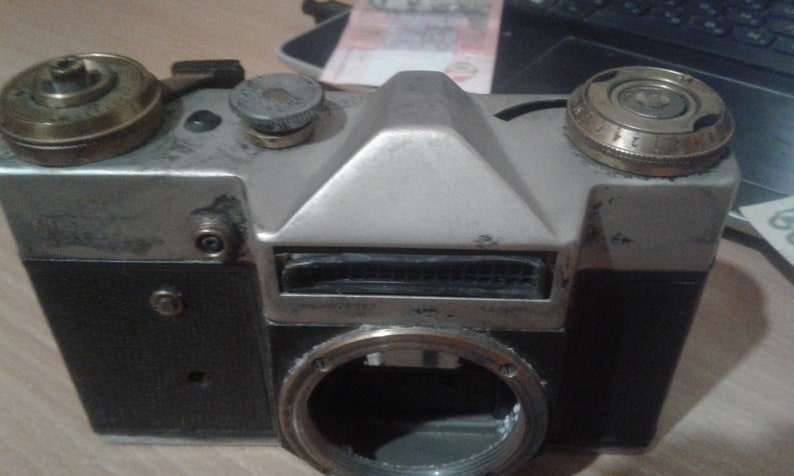 Puede incluir: Una c&aacute;mara de pel&iacute;cula vintage de 35 mm de color plateado y negro con una lente de lat&oacute;n y una correa de cuero negra. La c&aacute;mara tiene una parte superior plateada y una parte inferior negra con un anillo de lente plateado. La c&aacute;mara tiene una correa de cuero negra unida al lateral.