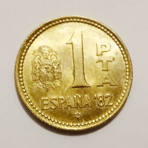 Puede incluir: Una moneda española de color dorado con el año 1982 y el texto "ESPAÑA" en el reverso. El anverso de la moneda presenta el escudo de armas español y el número "1" con la letra "P" de "Peseta" a su lado.