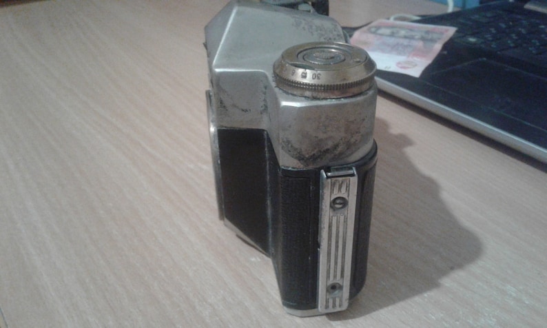 Puede incluir: Una c&aacute;mara de pel&iacute;cula vintage plateada y negra con un acabado desgastado. La c&aacute;mara tiene una cubierta de cuero negro y un cuerpo de metal plateado. La c&aacute;mara tiene un dial en la parte superior con los n&uacute;meros del 1 al 30.