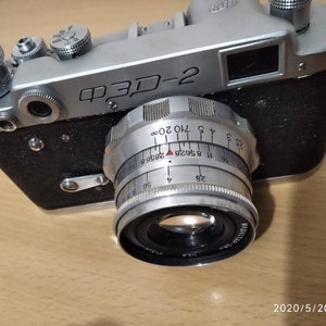 Puede incluir: Una cámara de película de 35 mm de color plateado y negro con una lente marcada con "FED-2" y una escala de profundidad de campo. La cámara tiene una cubierta de cuero negro y un cuerpo de metal plateado.