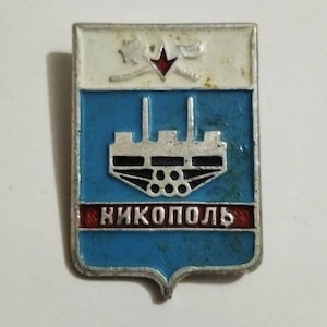 Pode incluir: Um alfinete retangular com um design de escudo. A seção superior é branca com uma estrela vermelha e um design. A seção do meio é azul com uma estrutura prateada. A seção inferior é vermelha com a palavra "НИКОПОЛЬ" em branco.