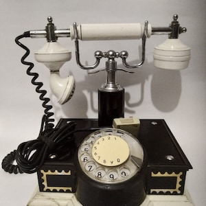 Könnte beinhalten: Ein Vintage-Drehwahltelefon mit schwarzem Sockel und weißen Akzenten. Das Telefon hat ein weißes Drehwahlrad mit schwarzen Zahlen und ein schwarzes Hörer mit einem weißen Hörer. Das Telefon befindet sich auf einer weißen Oberfläche.