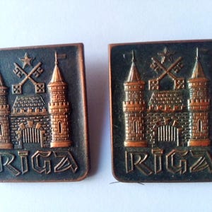 May include: Two vintage copper-colored metal pins with the coat of arms of Riga, Latvia. The pins feature a stylized castle with two towers and a gate, with crossed keys and a star above. The word "RIGA" is inscribed below the castle.