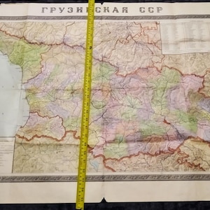 May include: Vintage map of the Georgian Soviet Socialist Republic, featuring detailed geographical information. The map is multi-colored, with a yellow measuring tape laid across it. The map is approximately 60 inches long.
