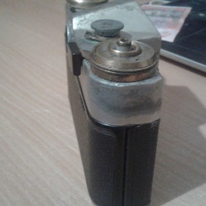 Puede incluir: Una c&aacute;mara vintage de plata y negro con una lente de lat&oacute;n y una cubierta de cuero negro. La c&aacute;mara tiene una superficie texturizada y una peque&ntilde;a perilla en la parte superior.