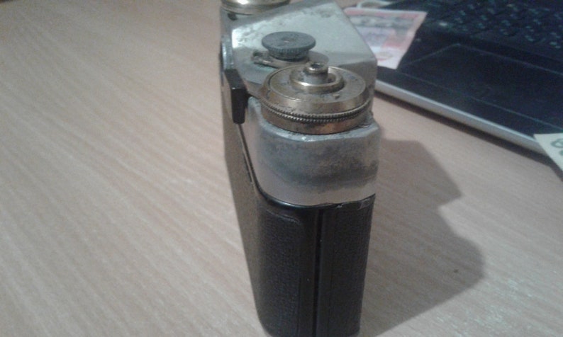 Puede incluir: Una c&aacute;mara vintage de plata y negro con una lente de lat&oacute;n y una cubierta de cuero negro. La c&aacute;mara tiene una superficie texturizada y una peque&ntilde;a perilla en la parte superior.