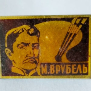 Op de afbeelding: Een rechthoekige, vintage luciferdoos met een portret van een man met een snor en de naam "M. VRUBEL" in gouden letters. De achtergrond bevat een palet en penselen, wat wijst op een kunstgerelateerd thema.