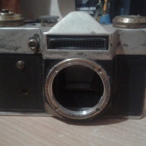 Puede incluir: Una c&aacute;mara de pel&iacute;cula vintage de 35 mm de color plateado y negro con un anillo de lente plateado y una cubierta de cuero negro. La c&aacute;mara tiene una placa superior plateada con una ventana de contador de pel&iacute;cula negra.