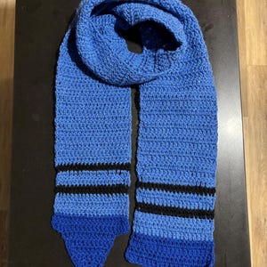 May include: A handmade blue crochet scarf with black stripes. The scarf is made of a soft, textured yarn and features a solid blue color with two black stripes near the bottom. The scarf is approximately 70 inches long.