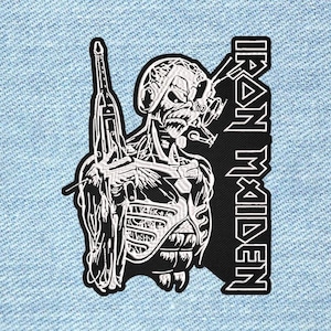 May include: A black and white embroidered patch featuring a skeletal figure wearing a headset and holding a rifle, positioned next to the text IRON MAIDEN in a bold, stylised font. The patch is applied to a light blue denim fabric background.
