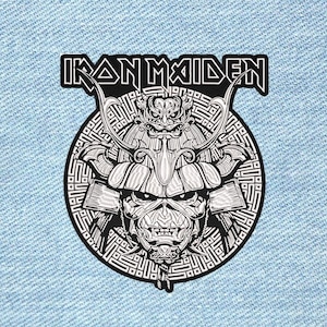 May include: A black and white embroidered patch featuring the text IRON MAIDEN above a circular design. The graphic depicts a stylised character wearing a detailed samurai helmet with horns, set against a background of intricate geometric patterns.