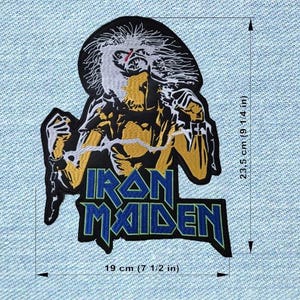 Op de afbeelding: Een geborduurde Iron Maiden patch met de mascotte van de band in zwart, wit en goudkleurig garen. Het ontwerp bevat de tekst Iron Maiden in het blauw. De afmetingen zijn 23,5 cm bij 19 cm, zoals aangegeven door de zichtbare meetlijnen.