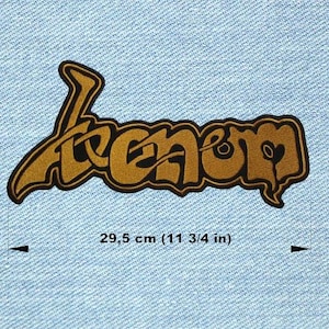 Könnte beinhalten: Ein gold-schwarzer, gestickter Aufnäher mit dem Wort "Venom" in einer stilisierten Schriftart. Der Aufnäher ist 29,5 cm (11 3/4 Zoll) lang und vor einem hellblauen Jeanshintergrund platziert. Der Aufnäher hat einen schwarzen Rand.