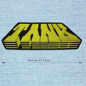 Könnte beinhalten: Ein gelb-schwarzer Aufnäher mit dem Wort "TANK" in einer stilisierten 3D-Schriftart. Der Aufnäher ist etwa 29,5 cm lang und befindet sich auf einem hellblauen Jeans-Hintergrund. Der Aufnäher hat einen schwarzen Rand.