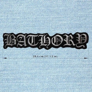 May include: A black and white embroidered patch with the word "BATHORY" in gothic lettering. The patch is 11 1/2 inches long and is set against a light blue denim background. The patch has a black border.
