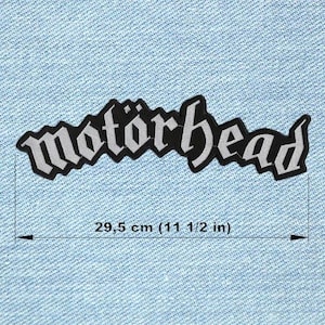 Pode incluir: Um patch bordado em preto e branco com a palavra "Motörhead" em uma fonte estilizada. O patch tem aproximadamente 29,5 cm de comprimento e está sobre um fundo de ganga azul clara.